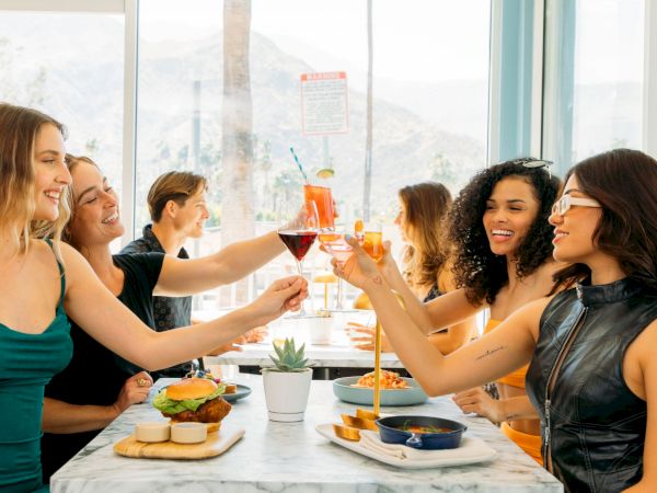 A group of friends toasts drinks at a restaurant table with food, enjoying a sunny day.