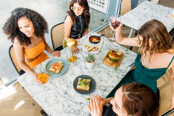 Four women enjoy food and drinks at a marble table, featuring pizza and a burger, in a stylish setting.