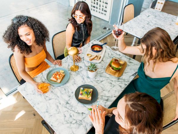 Four women enjoy food and drinks at a marble table, featuring pizza and a burger, in a stylish setting.