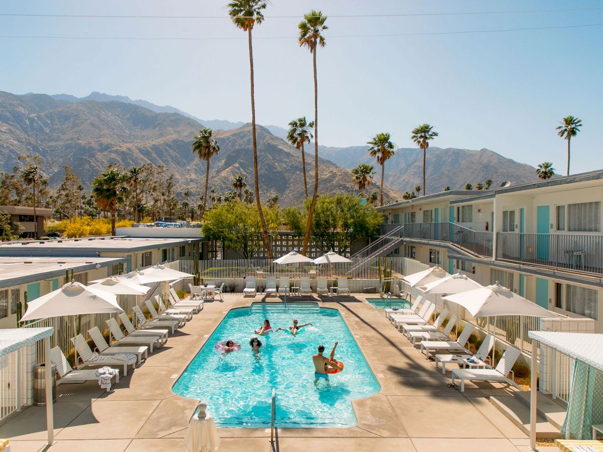 A sunny hotel pool surrounded by loungers and umbrellas, with people swimming and palm trees in the background, set against mountain views.