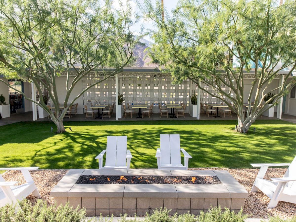 The image shows a serene backyard with white chairs around a fire pit, lush green grass, and trees providing shade and privacy to a sitting area.