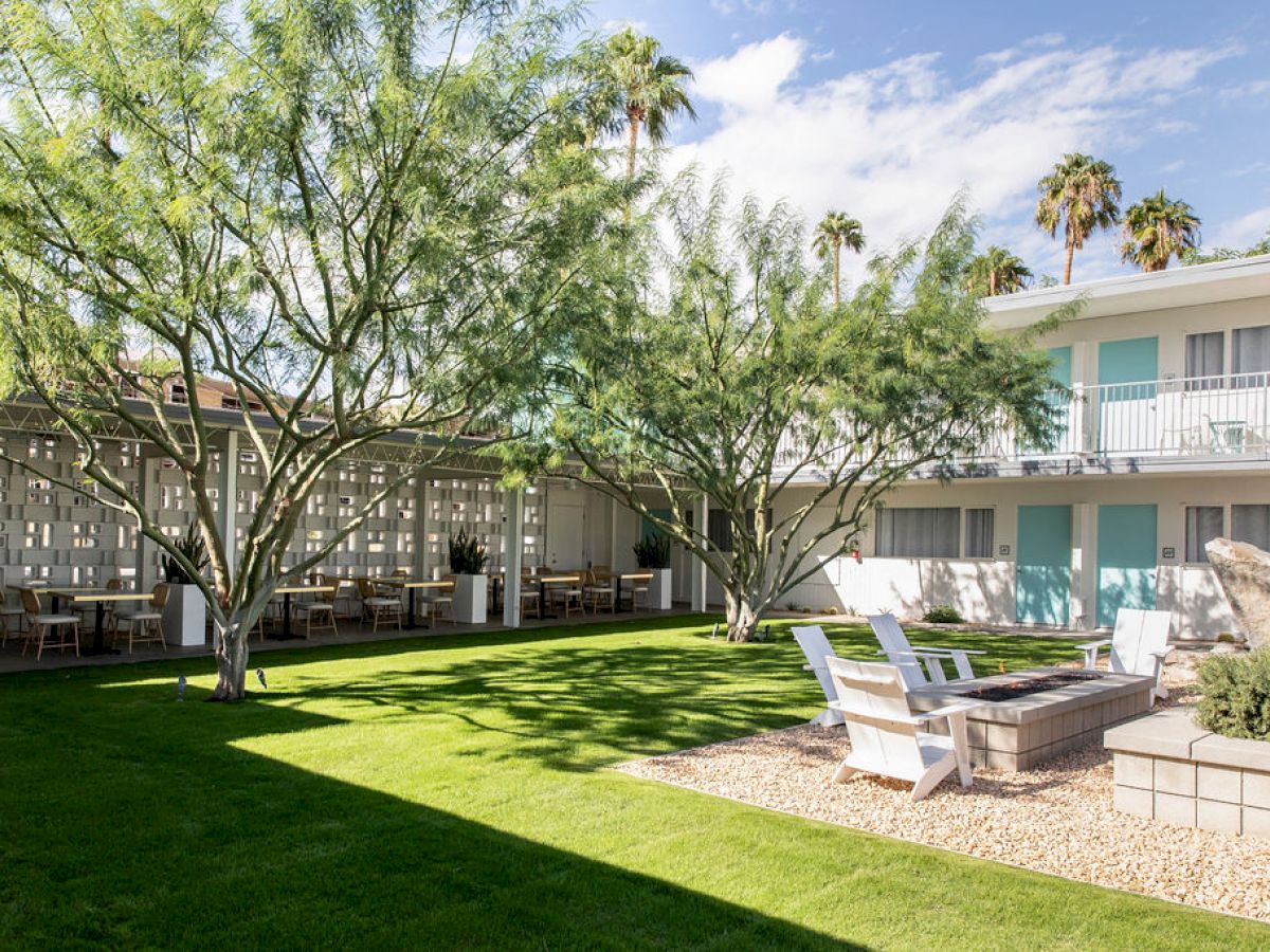 A sunny courtyard with lush greenery, seating areas, and a modern building in the background, under a bright blue sky.