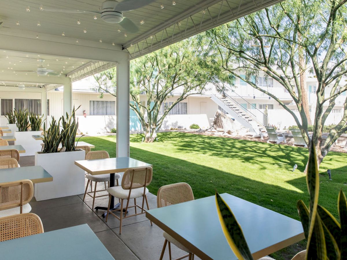 An outdoor seating area features tables and chairs, surrounded by greenery and string lights, with a view of a garden and building.