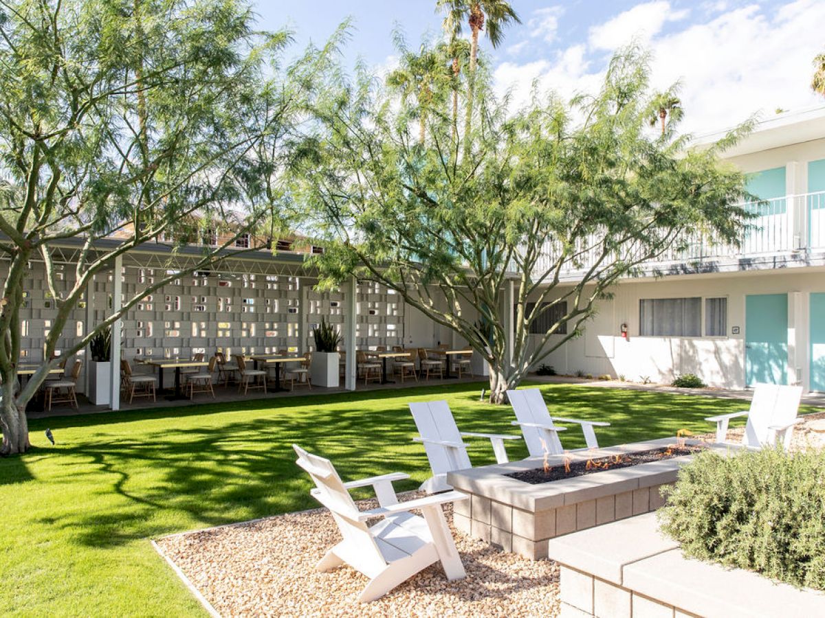 Outdoor seating area with white chairs around a fire pit, green lawn, trees, and a building with blue doors in the background.