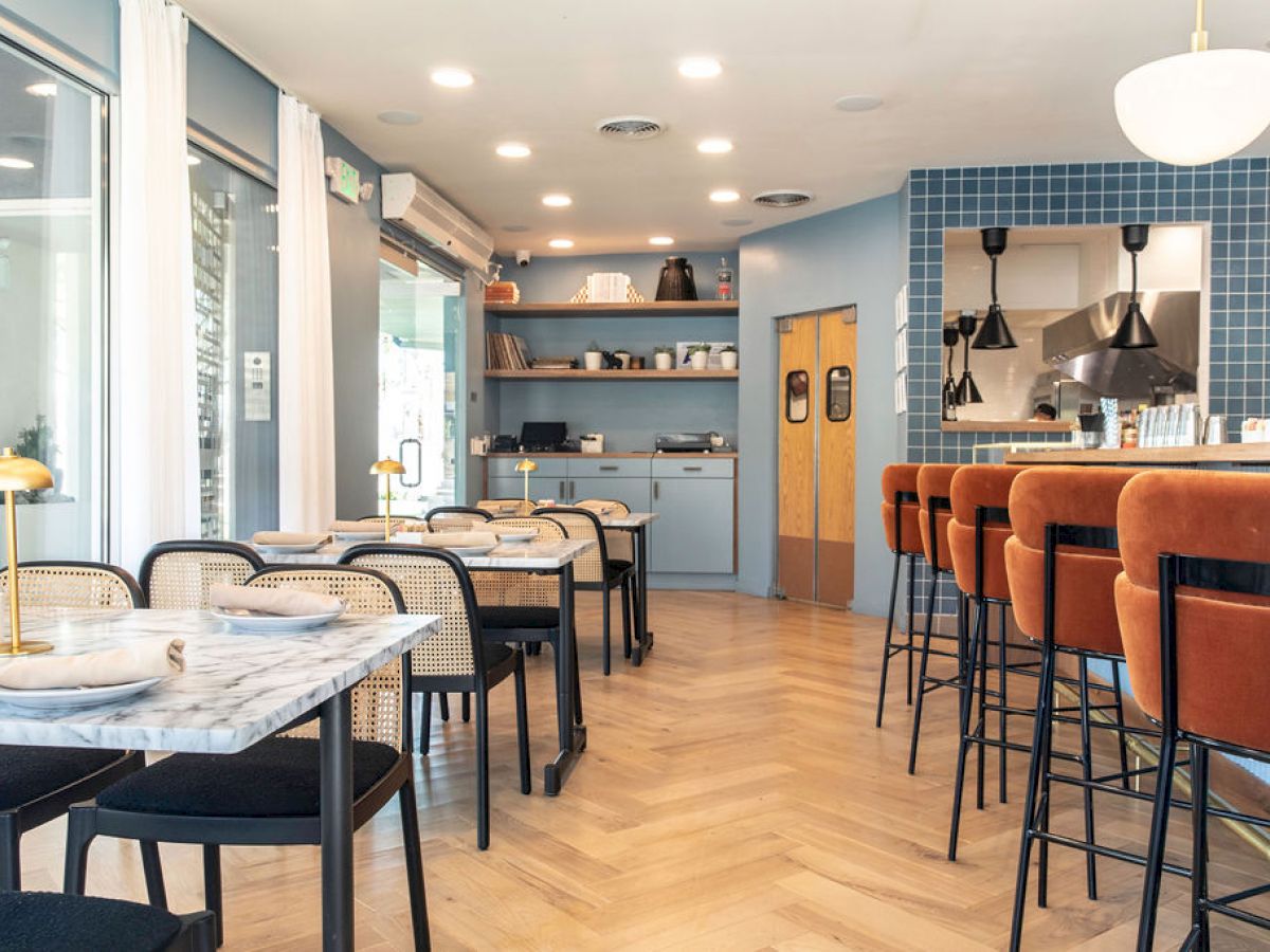 A modern restaurant interior with marble tables, black chairs, and a bar with orange stools, featuring light blue walls and wooden flooring.