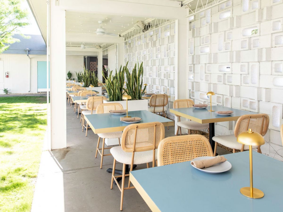 An outdoor dining area with blue tables, wicker chairs, and decorative plants under a covered patio with soft lighting.