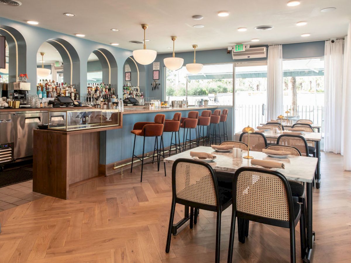 A stylish, modern restaurant interior with a bar, pendant lights, and seating with a view of the outdoors through large windows.