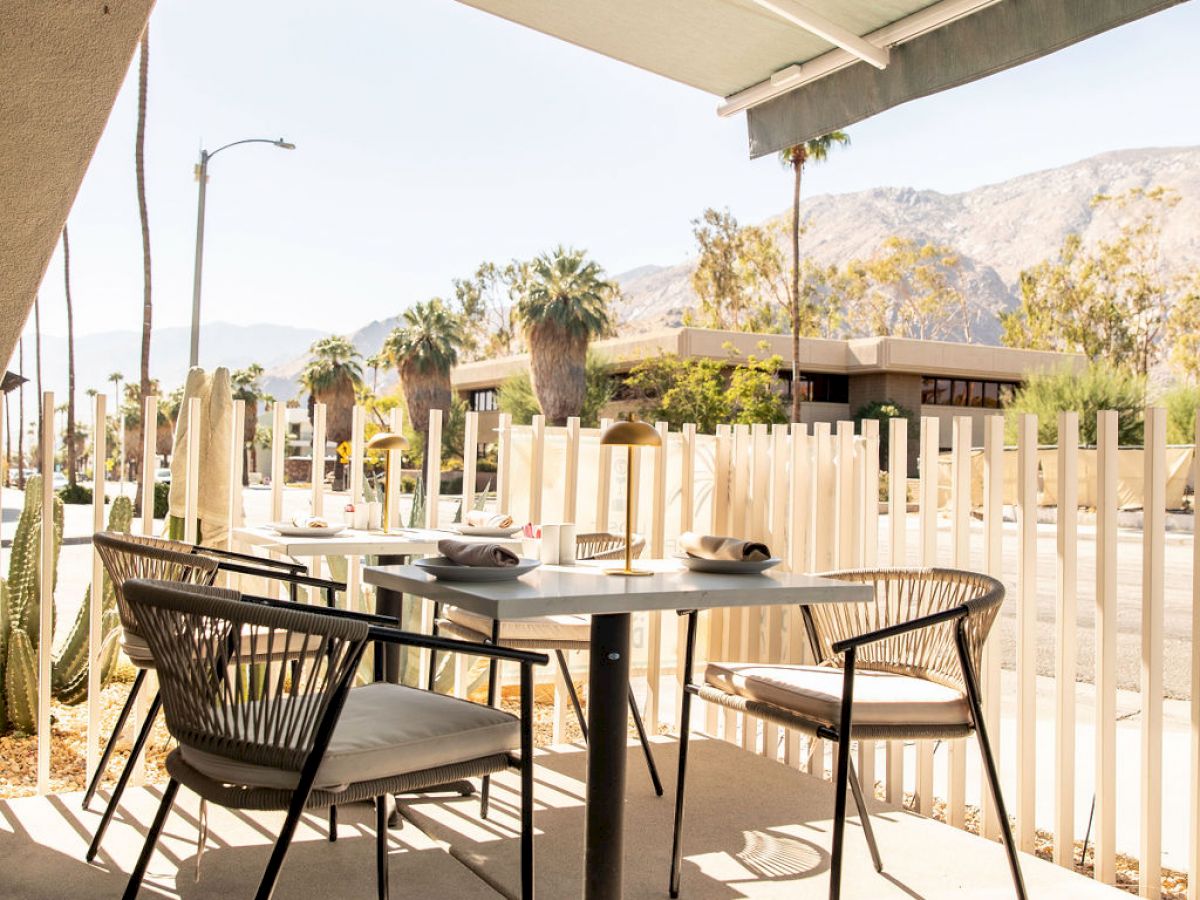 Outdoor patio with a table and chairs under an awning, overlooking a street with palm trees and mountains in the background.