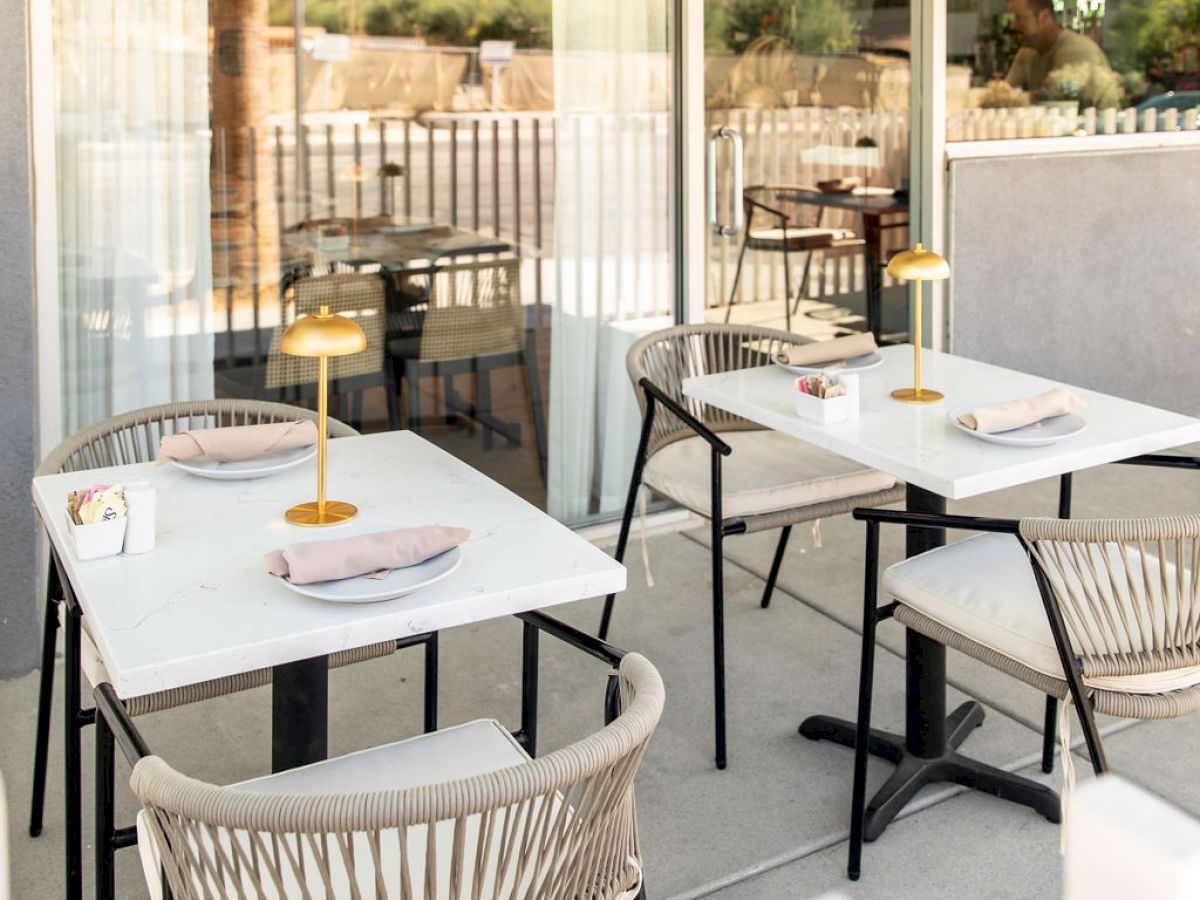 The image shows an outdoor patio area of a restaurant with neatly set tables and chairs, featuring small lamps and a modern design.