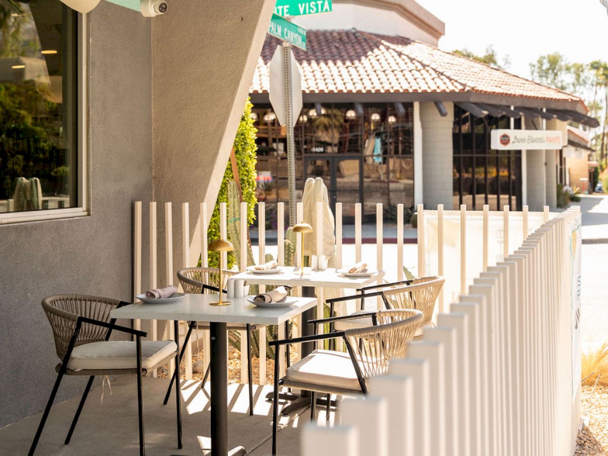 An outdoor cafe patio with a small table, chairs, and white picket fence; nearby a street and building are visible.