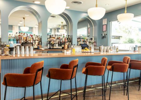 The image shows a stylish bar with orange chairs, a well-stocked counter, large mirrors, and hanging globe lights in a bright, modern setting.