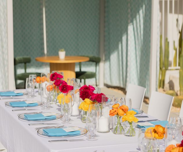 An elegantly set outdoor dining table with blue napkins and colorful floral centerpieces, under a shaded pergola with a cozy seating area.