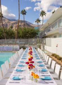 An outdoor dining setup by a pool, with a long table lined with white chairs, colorful flowers, and blue napkins, surrounded by a scenic view.