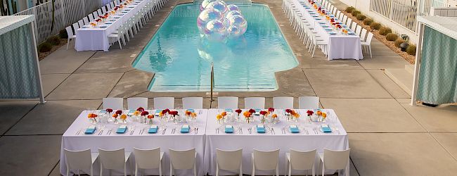 A poolside scene with elegant dining tables set up, featuring clear inflatable balls in the pool, surrounded by a hotel-like building.