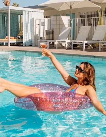 A person relaxes on a colorful float in a pool, holding a drink. Lounge chairs and umbrellas are in the background.