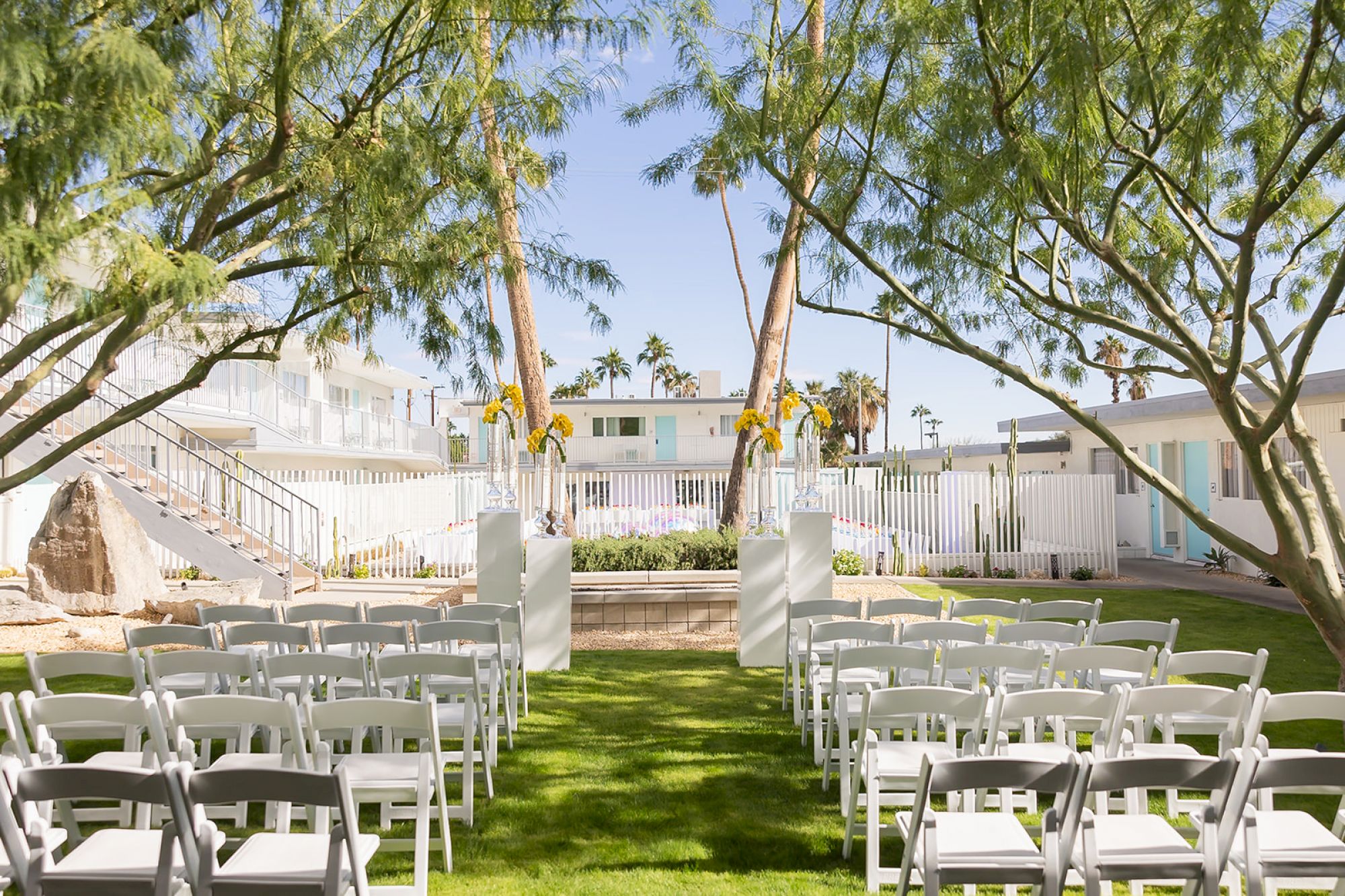 An outdoor wedding setup with white chairs arranged on a grassy lawn, flanked by trees, facing a small altar.