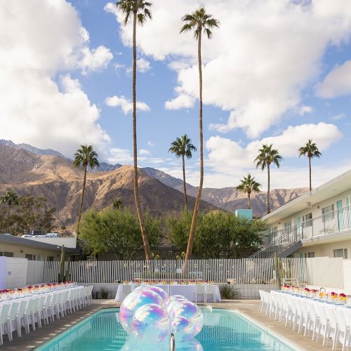 An outdoor setting with a pool, balloons, and two long tables set for an event; mountains and palm trees in the background.