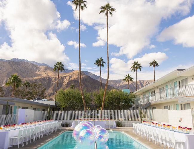 An outdoor setting with a pool, balloons, and two long tables set for an event; mountains and palm trees in the background.