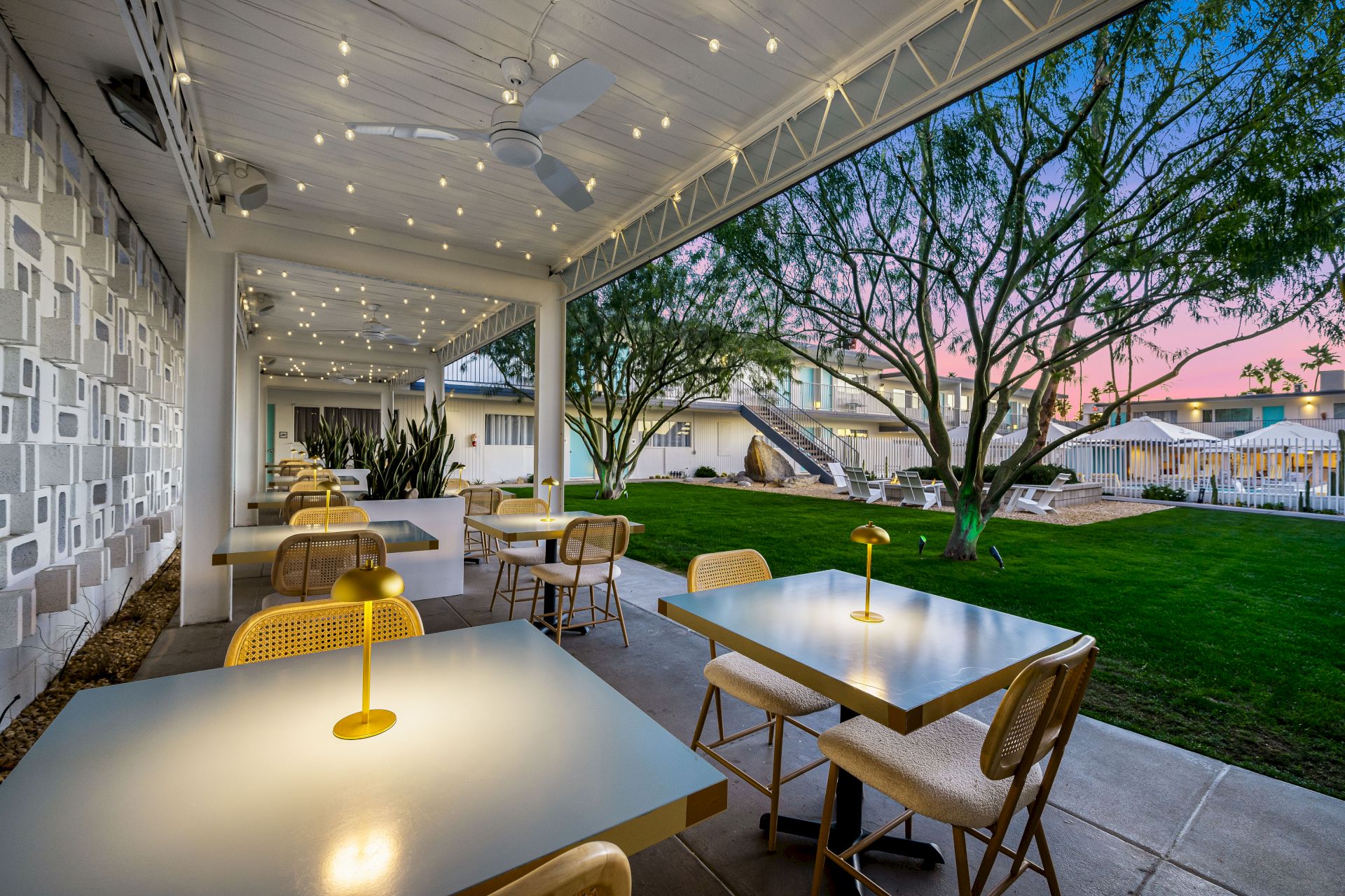 Outdoor dining area with tables, chairs, and warm lighting under string lights at sunset. Lush garden and buildings in the background.