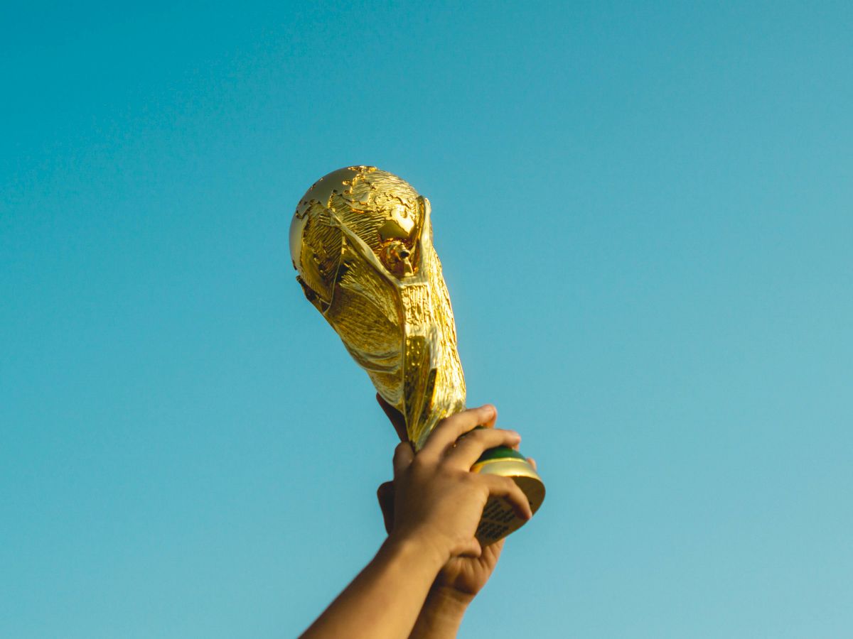 A hand lifts a gold soccer/football trophy against a clear blue sky, celebrating victory.