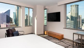 This image depicts a modern hotel room with a bed, a TV, a chair, a small table, and large windows with views of a cityscape.