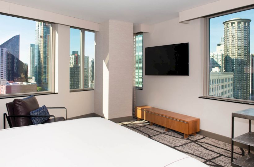 This image depicts a modern hotel room with a bed, a TV, a chair, a small table, and large windows with views of a cityscape.