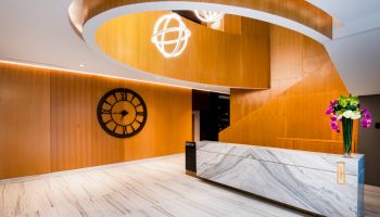 A modern lobby with a large wall clock, curved wooden ceiling, marble reception desk, and floral arrangement on the right side.