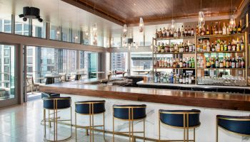 A modern bar with a wooden ceiling, blue and gold barstools, and a well-stocked liquor shelf, featuring floor-to-ceiling windows and city views.