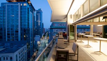 A modern cityscape viewed from a balcony at dusk, featuring high-rise buildings and a stylish outdoor seating area along with indoor tables.