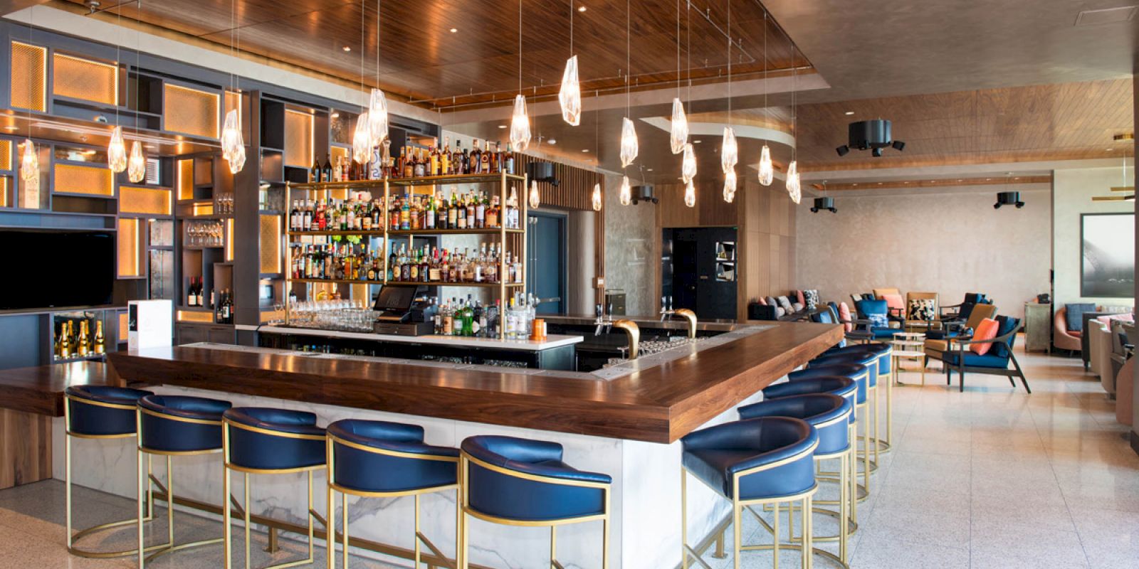 A modern bar with a central counter, blue barstools, hanging pendant lights, and a well-stocked liquor shelf, surrounded by comfortable seating areas.