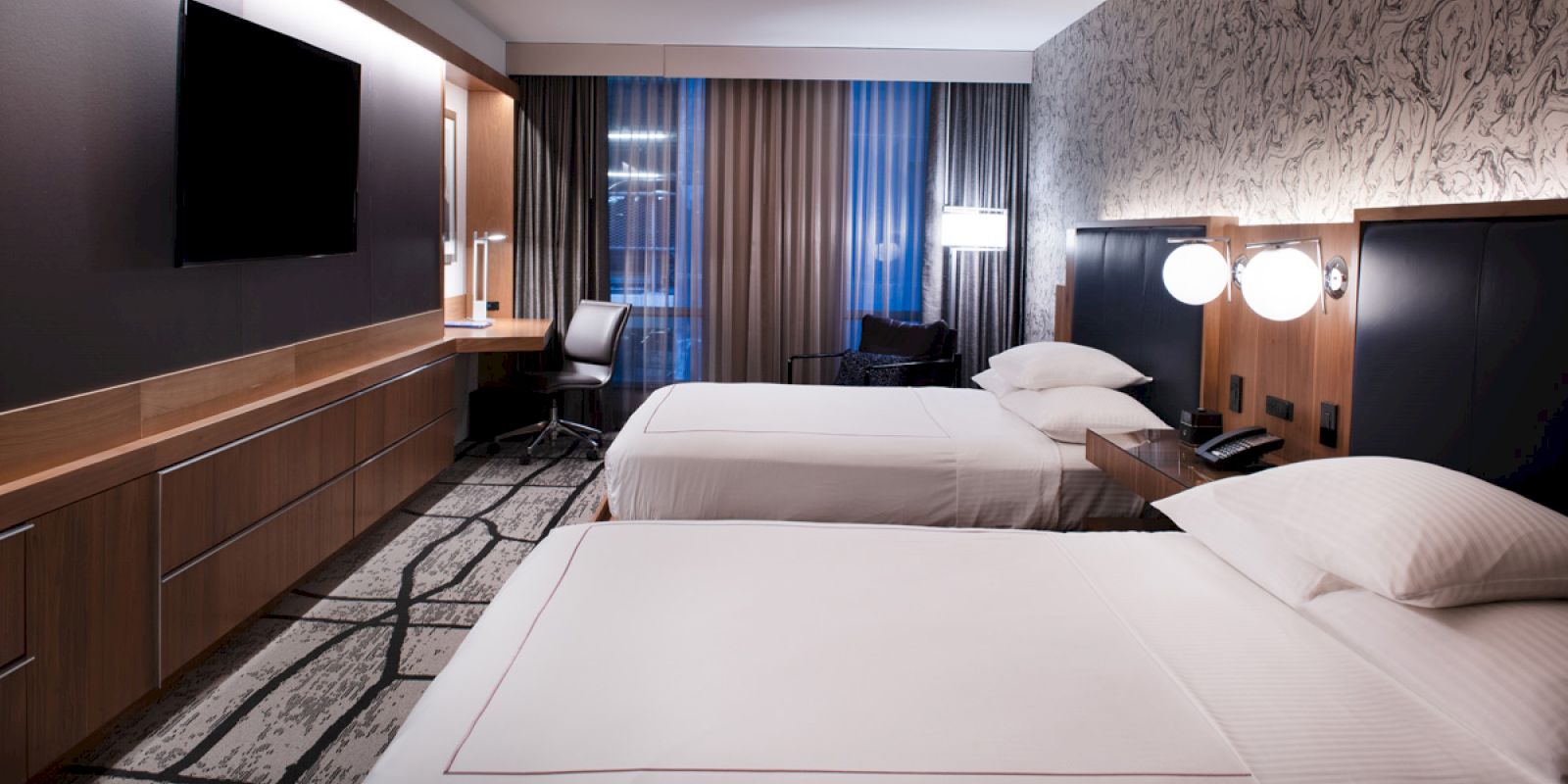 A modern hotel room with two neatly made beds, a wall-mounted TV, a work desk with a chair, and a patterned carpet, all in a stylish setting.