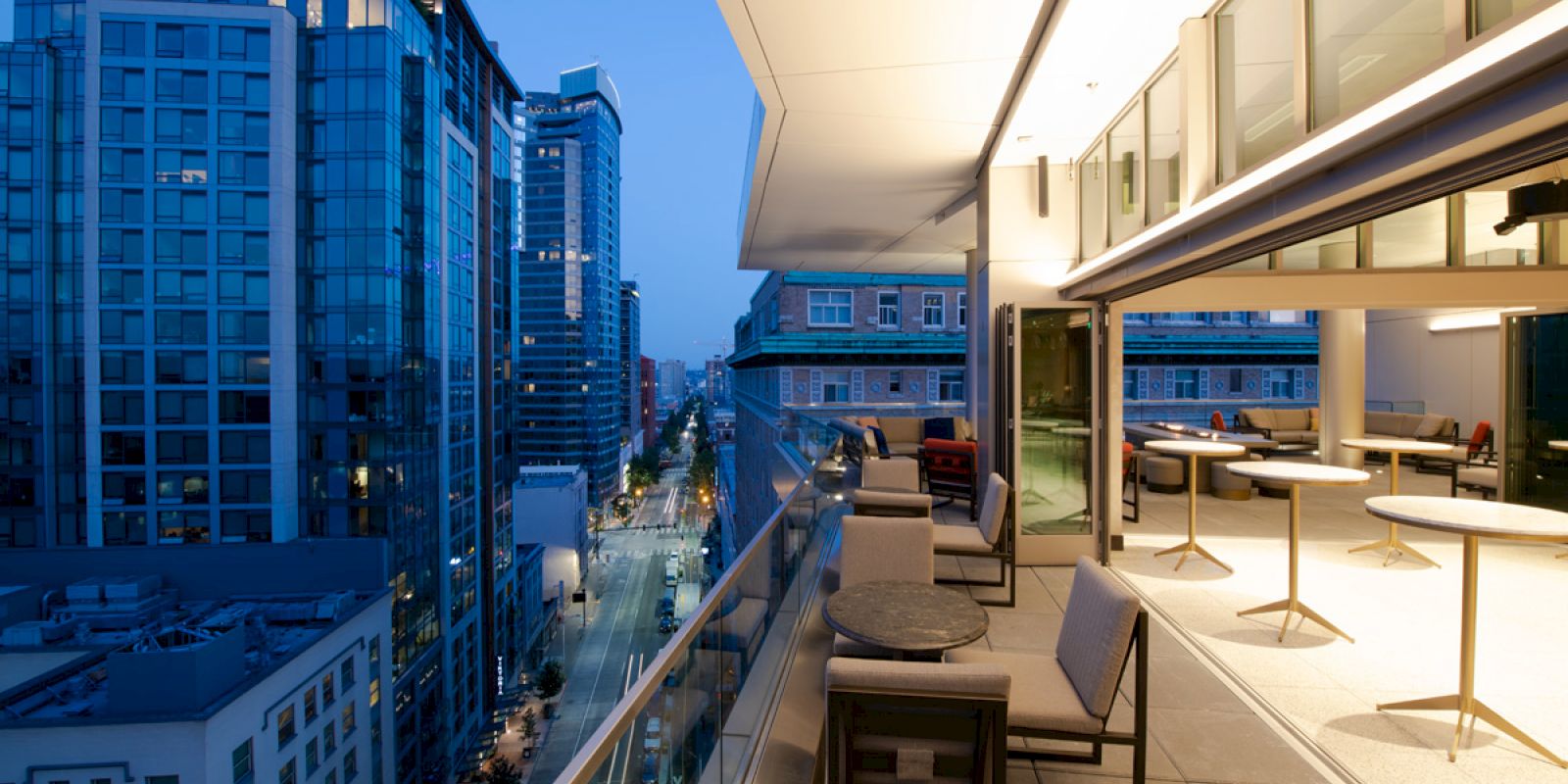 A modern urban balcony at dusk, equipped with tables and chairs, overlooks a street lined with high-rise buildings.