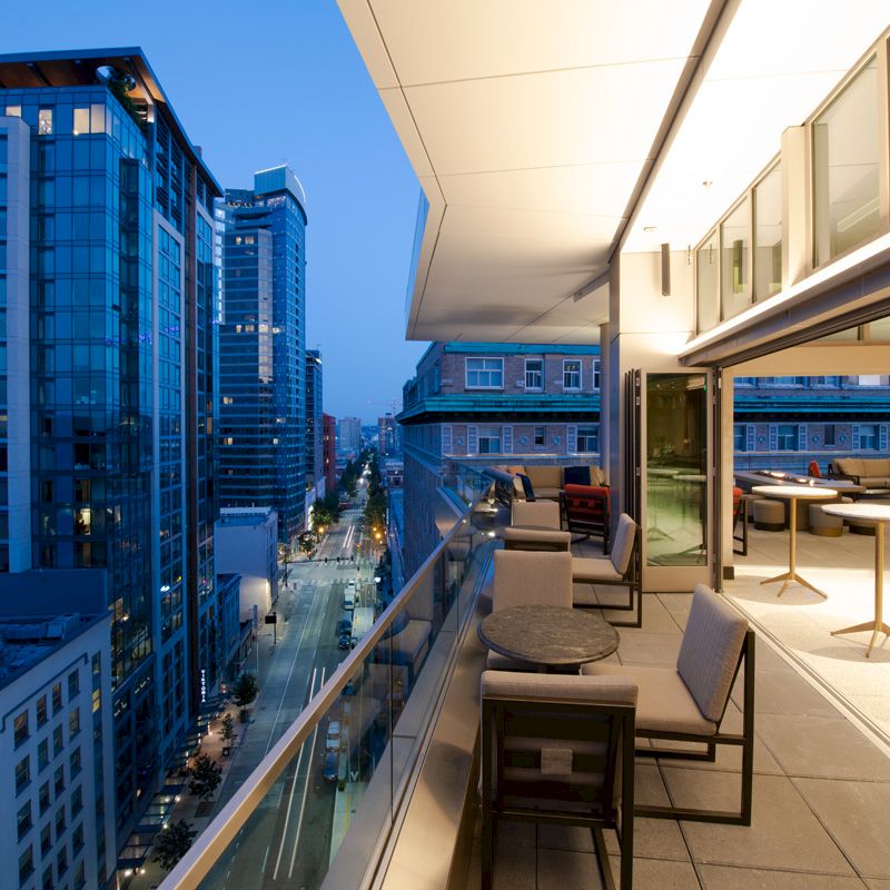 A modern outdoor balcony space with seating and tables overlooks a city street lined with tall buildings at dusk.