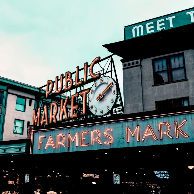 The image shows an iconic "Public Market" sign with a clock and a neon "Farmers Market" sign beneath it, likely at Pike Place Market in Seattle.