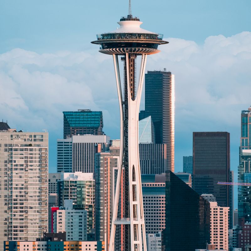 The image shows the Space Needle, a famous observation tower surrounded by modern skyscrapers against a blue sky backdrop.