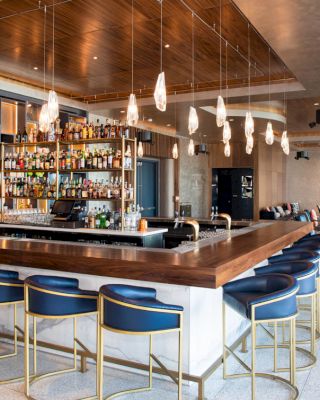 The image shows a modern bar with a wooden countertop, blue cushioned stools, hanging lights, and a well-stocked bar in the background.