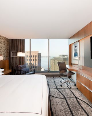 A modern hotel room with a large bed, wall-mounted TV, desk, chair, and a window offering a cityscape view.