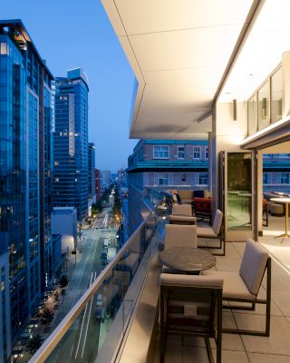 A modern rooftop terrace in a city with seating areas overlooks a street lined with high-rise buildings during the evening.