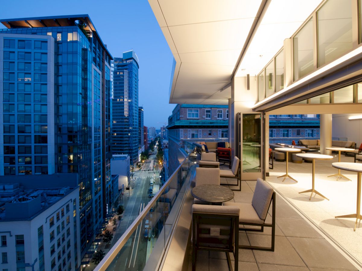 A modern cityscape at dusk is viewed from a high-rise balcony with outdoor seating and tables. The glass buildings reflect the evening sky.