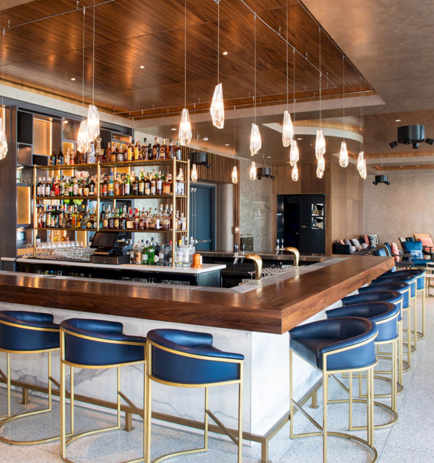 A modern bar with a wooden countertop, blue chairs, a wide selection of bottles, pendant lights, and seating areas in the background.