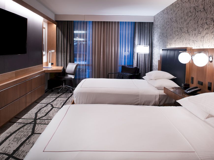 Modern hotel room with two double beds, wall-mounted TV, desk with chair, nightstand, and contemporary decor.
