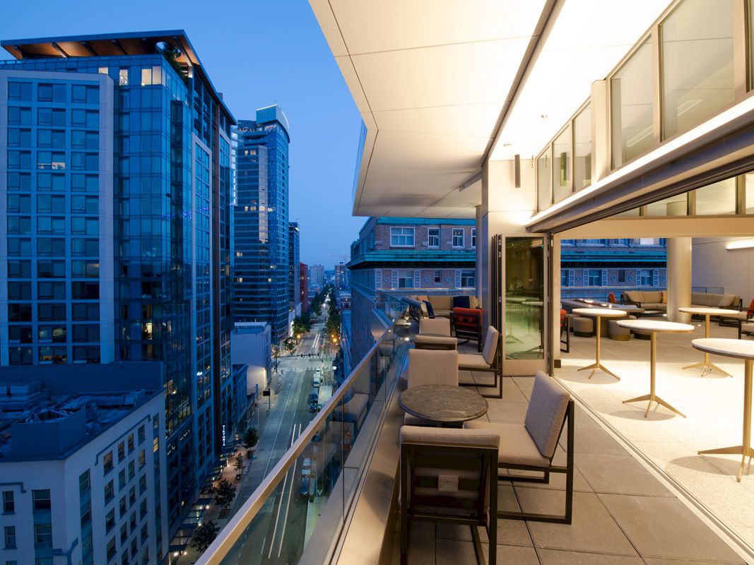 A modern cityscape at dusk viewed from a balcony with outdoor seating and adjacent indoor dining areas illuminated by warm lighting.