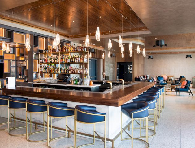 A modern bar with a wooden counter, blue stools, hanging lights, and a well-stocked shelf of liquor bottles, with seating areas in the background.