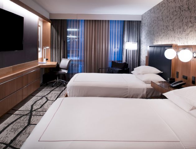 A modern hotel room with two double beds, a wall-mounted TV, a desk with a chair, and contemporary decor, including patterned wallpaper and a large window.