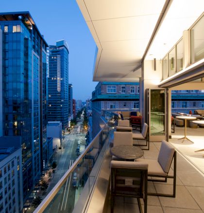 A modern urban balcony at dusk with outdoor seating and tables overlooks a cityscape with tall buildings and a street.