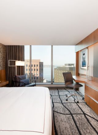 The image depicts a modern hotel room with a large bed, wall-mounted TV, desk, chair, and a window offering a city view.