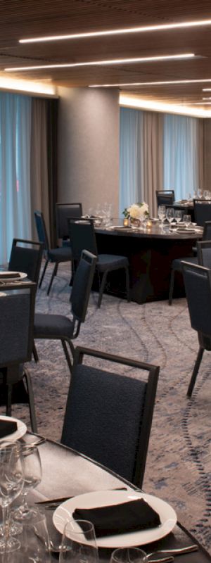 The image shows a sophisticated banquet hall with round tables set for an event, featuring elegant table settings and modern decor with dim lighting.