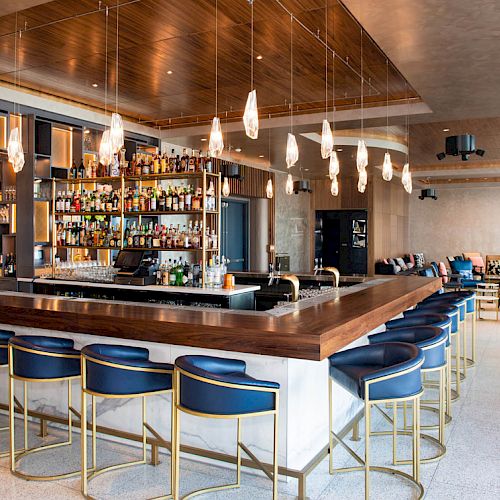 The image shows a modern bar with a wooden counter, blue bar stools, hanging lights, and a well-stocked liquor shelf in a stylish, open space.