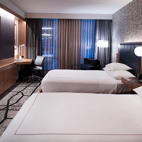 The image shows a modern hotel room with two beds, a wall-mounted TV, a desk with a chair, and stylish lighting and decor.