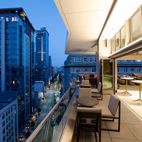 The image shows a modern urban balcony with seating, tables, and views of high-rise buildings at dusk. The interior is visible with more seating areas.